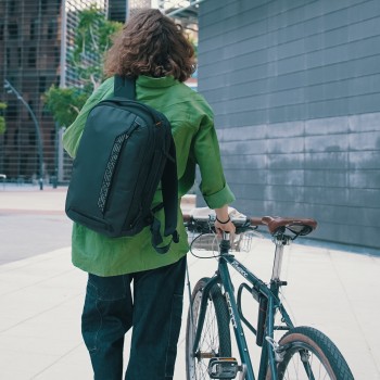 Targus Commuter EcoSmart 16" backpack, stylish in black for urban environments A person walking in a green jacket with a black Targus backpack on their back, next to a bicycle and with urban buildings in the background.